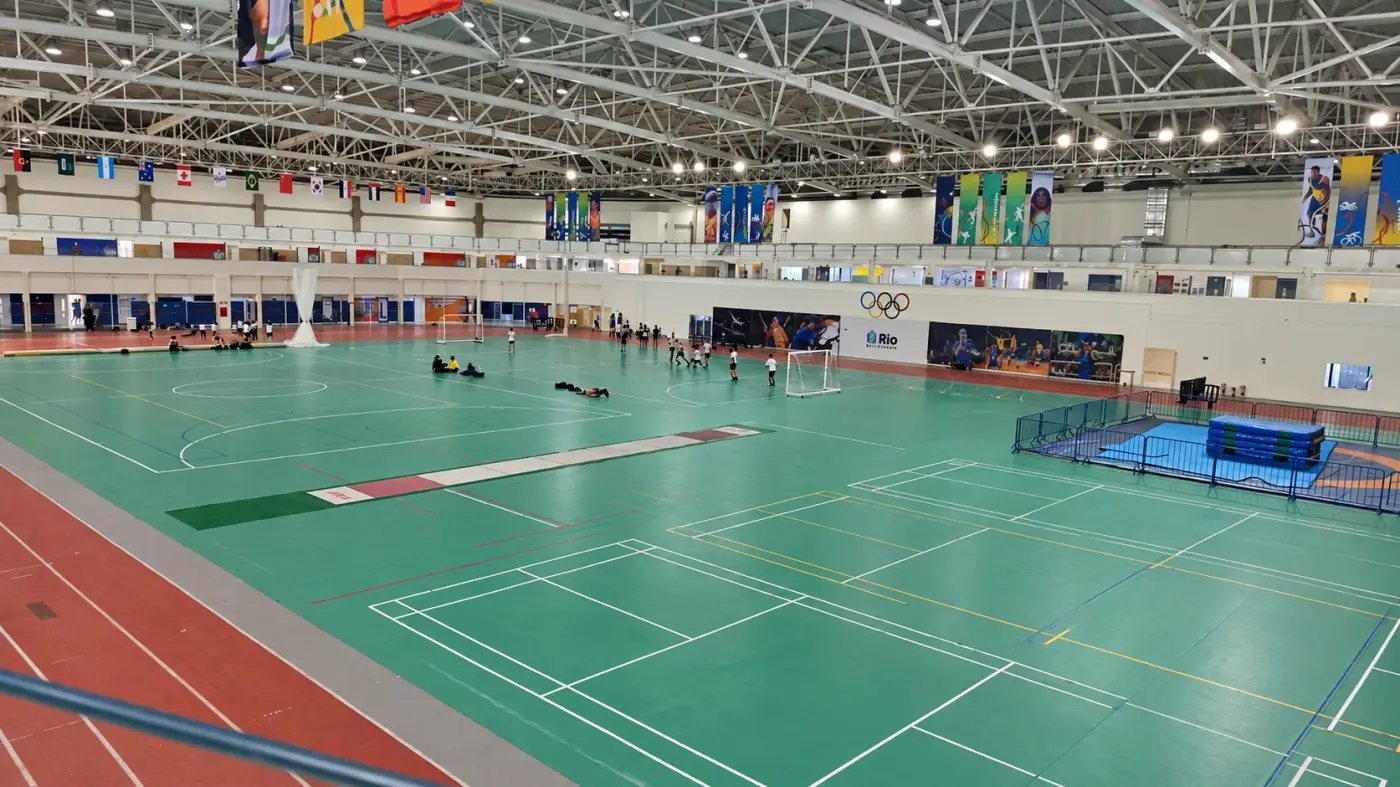 O ginásio visto da arquibancada: quadras verdes, pista de atletismo e bandeiras dos países no teto.